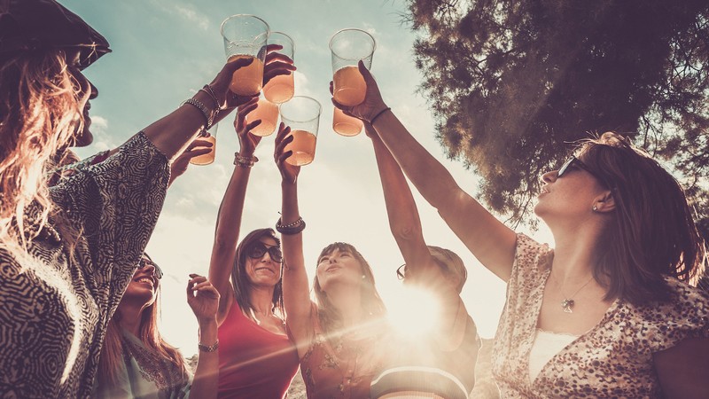 celebrating friendship or success team works outdoor with colored juice and 7 beautiful females friends all together with orange cocktail and the sunlight near the beach and the ocean. nice group of people summer style
