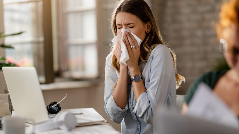 Young sick businesswoman sneezing in a tissue while working in the office.